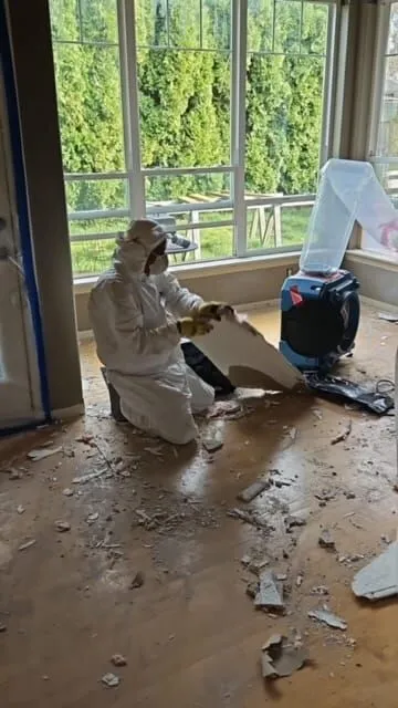 A person in protective gear carefully removes drywall in a room featuring a large window, almost like prepping an art gallery space. Debris is scattered across the floor, while a blue air scrubber hums nearby, adding to the setting of this unconventional contemporary art exhibition.