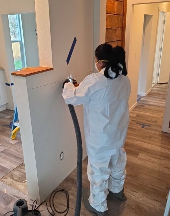 Person in white protective suit and mask uses a tool with a hose to cut or sand a wall during home renovation. Blue painter's tape marks areas on the walls and floors inside the house under renovation.
