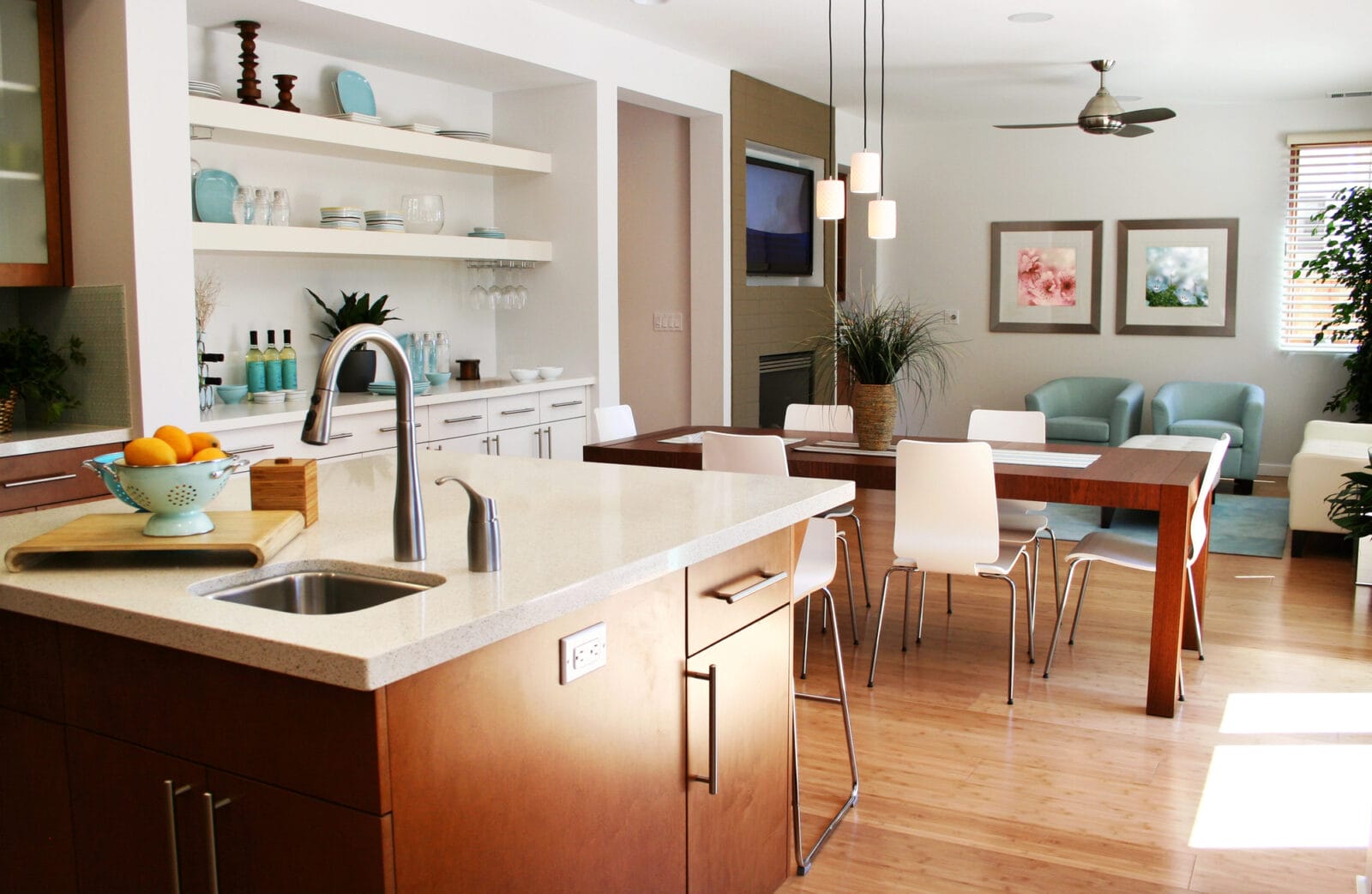 Modern open-plan kitchen and dining area with wooden flooring, a central island, white chairs, and decor in soft tones. A sitting area with plants is visible in the background, meticulously maintained by professional house cleaning services.