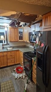 A kitchen with wooden cabinets and appliances resembles a chaotic gallery, highlighted by a partially collapsed ceiling and scattered debris on the floor. A red bucket and broom stand nearby, adding to the visual narrative of disarray.