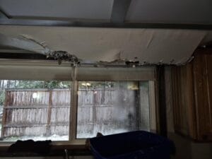 A damaged ceiling with water dripping through, located near a window with a gallery view of a wooden fence outside. A blue container is placed below to catch the water.