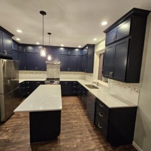 Modern kitchen gallery featuring dark blue cabinets, white countertops, stainless steel appliances, and wooden flooring. An island sits at the center with pendant lighting above, offering a sleek and contemporary showcase for culinary delights.