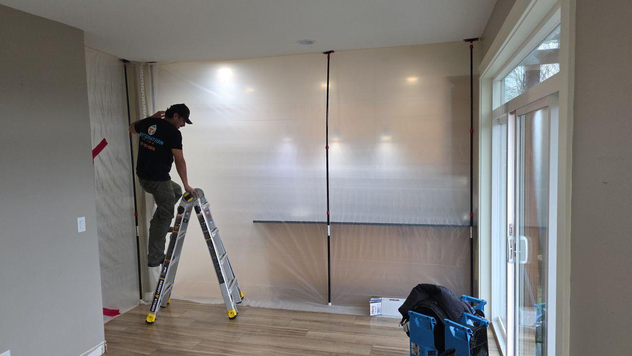 A person stands on a ladder securing plastic sheeting to section off part of a room for interior remodeling, with tools and equipment nearby on a wooden floor.