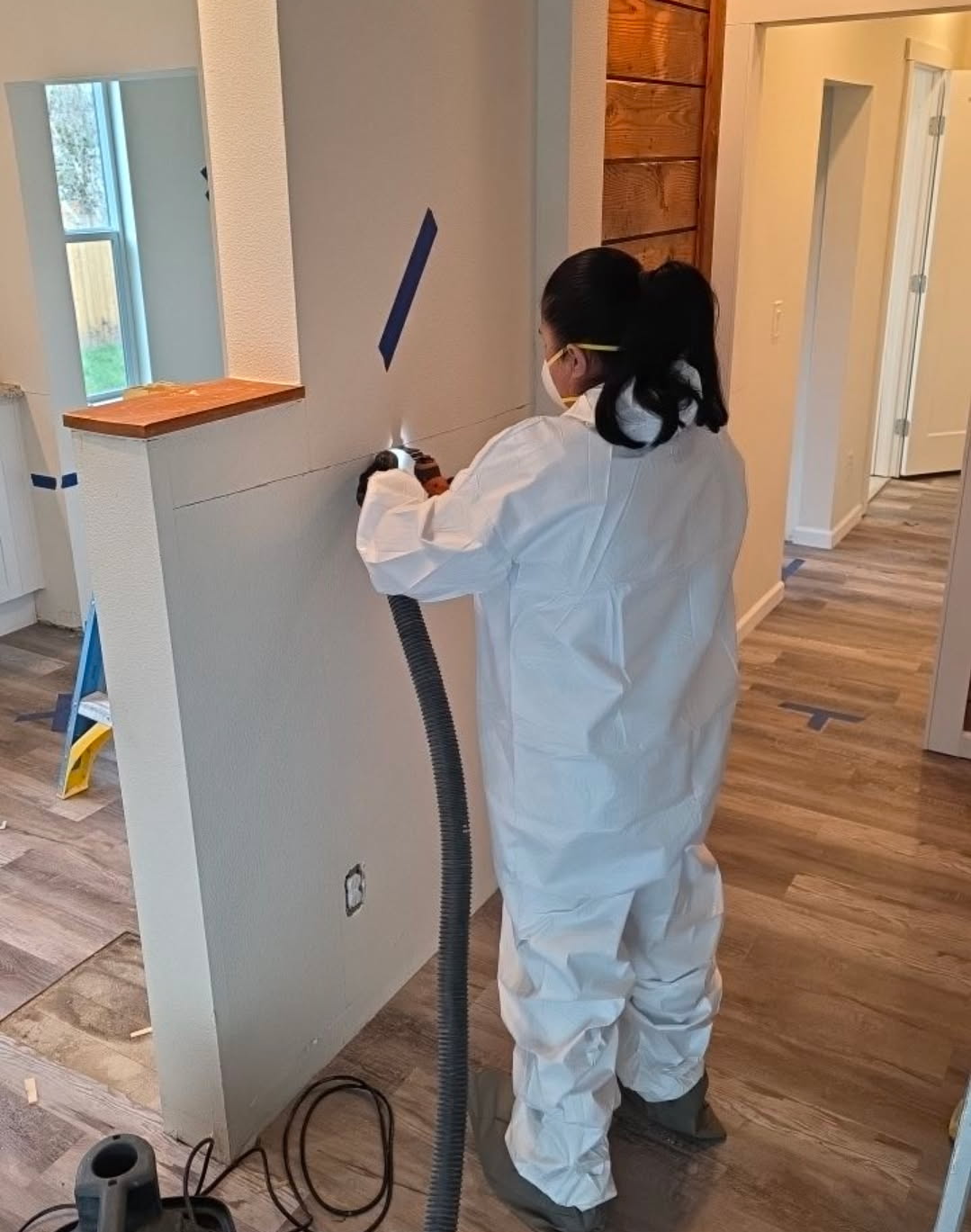 Person in white protective suit and mask uses a tool with a hose to cut or sand a wall during home renovation. Blue painter's tape marks areas on the walls and floors inside the house under renovation.