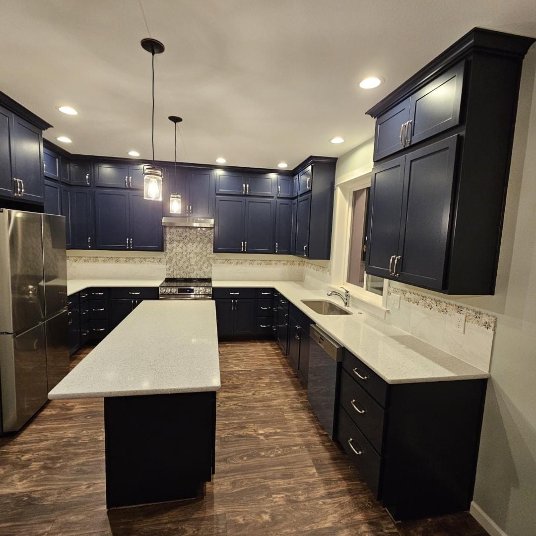 Modern kitchen with dark blue cabinets, stainless steel appliances, and a large island adorned with pendant lights. Wood flooring and stone countertops complete the design, making it an easy space to maintain with professional house cleaning services.